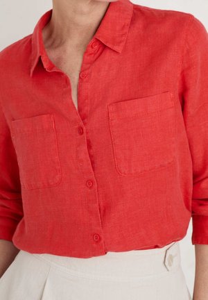 Woman wearing a red button-up shirt with two chest pockets and cream high-waisted pants, sleeves rolled up.