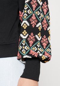 Sweatshirt preto com bordados florais e geométricos vibrantes em vermelho, amarelo, verde e branco nas mangas; punhos e bainha canelados.