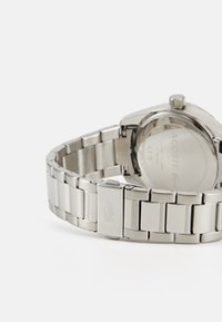 Ceas din oțel inoxidabil argintiu, cu carcasă rotundă și bratara integrată, având un închizător cu un logo de crocodil reliefat pe lateral.