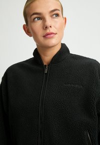 Giacca in pile nera con colletto alto, zip frontale e tasche laterali. Presenta una superficie strutturata e un marchio minimale.