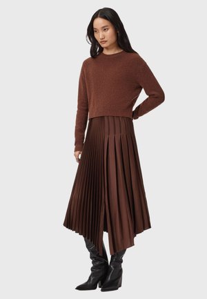Bruine cropped sweater van zachte textuur, gecombineerd met een lange, geplooide bruine rok. Het model draagt zwarte hakken.