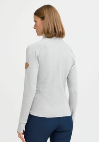 Lys grå langermet topp med høy hals, ribbestruktur og et lær-logo-patch på venstre erme; vist fra bak.