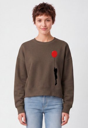 Femme portant un sweat-shirt marron avec une silhouette noire d'une personne tenant un ballon rouge imprimée sur la poitrine.
