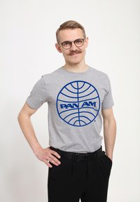 Šedé bavlněné tričko s velkým modrým kruhovým logem s textem "PAN AM". Modelka má krátké vlasy a brýle, tričko je kombinováno s černými kalhotami.