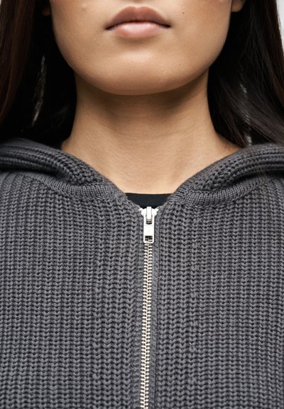 Partie inférieure du visage et cou d'une personne portant un sweat à capuche gris en maille avec une fermeture éclair argentée, cheveux foncés visibles sur les côtés.