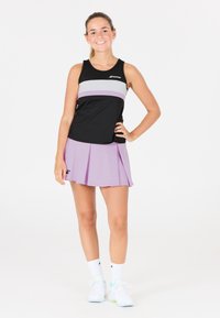 Giovane donna che indossa un top sportivo nero e lavanda abbinato a una gonna da tennis plissettata lavanda, calze bianche e scarpe da ginnastica bianche, sorridendo.