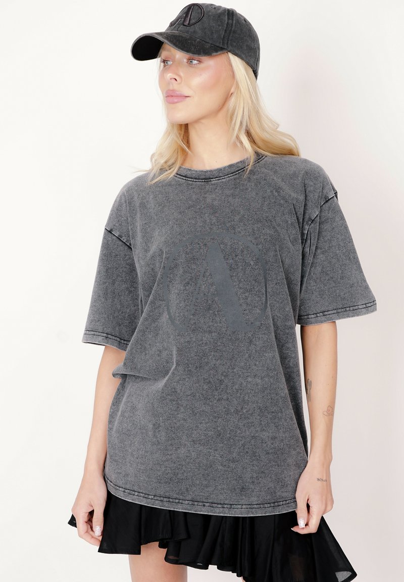 Szara, oversize koszulka z opuszczonymi ramionami, z okrągłym logo w ciemniejszym odcieniu. Materiał wydaje się miękki i ma fakturowane wykończenie.