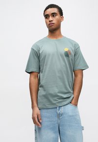 Gaiši zaļa kokvilnas īsroku T-krekls ar saulainu un viļņu dizainu oranžā, dzeltenā, zaļā un zilā krāsā kreisajā krūtī.