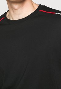 Sort T-shirt med rund hals, med røde accentlinjer på skuldrene og et lille hvidt logo på venstre side. Blødt, letvægtsstof.