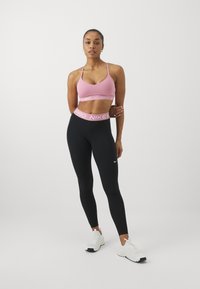 Sujetador deportivo rosa con logo, leggings negros con cinturilla en contraste y zapatillas deportivas blancas. La tela suave y el diseño ajustado acentúan la forma.