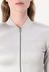 Světle šedá bomber bunda se zapínáním na zip, žebrovaným límcem a diskrétním logem na nápletu. Hladká, lesklá tkanina s přiléhavým střihem.