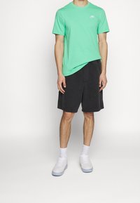 Muž v mátově zeleném tričku Nike, černých šortsích, bílých ponožkách a bílých teniskách stojí proti jednobarevnému bílému pozadí.