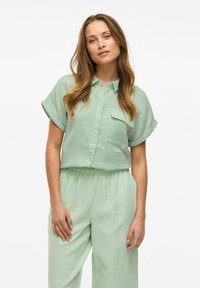 Camicetta verde chiaro a maniche corte con bottoni, dotata di una tasca sul petto, abbinata a pantaloni larghi dello stesso colore, entrambi in una texture stropicciata.