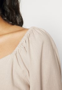 Beige linneblus med en samlad halsringning och puffärmar, med en slät yta och minimalistiska designelement.