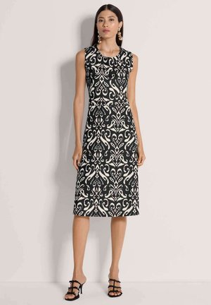 Frau trägt ein ärmelloses knielanges Kleid mit schwarz-weißem Muster, goldene Hängeohrringe und schwarze Riemchen-High Heels, steht vor einer hellen Wand.