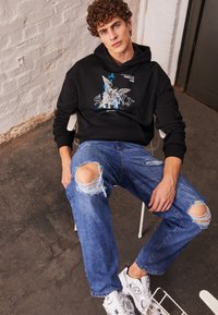 Sudadera negra con estampado gráfico y texto, combinada con jeans azules desgastados y zapatillas blancas. Materiales: mezcla de algodón, denim, goma.