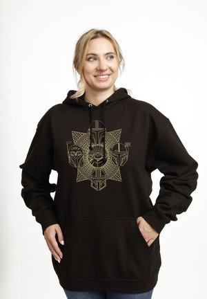Femme souriante portant un sweat à capuche noir avec un dessin en lignes dorées de cinq casques mandaloriens et un motif central représentant une créature, mains dans les poches.