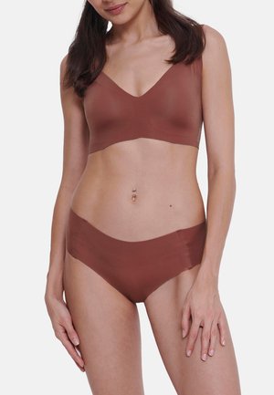 Kvinde med langt brunt hår iført sømløs brun bh og matchende hipster-underbukser, stående med armene afslappet mod hvid baggrund.