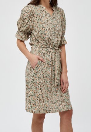 Femme portant une robe fleurie à manches courtes avec une ceinture nouée à la taille et une main dans la poche, debout devant un fond uni.