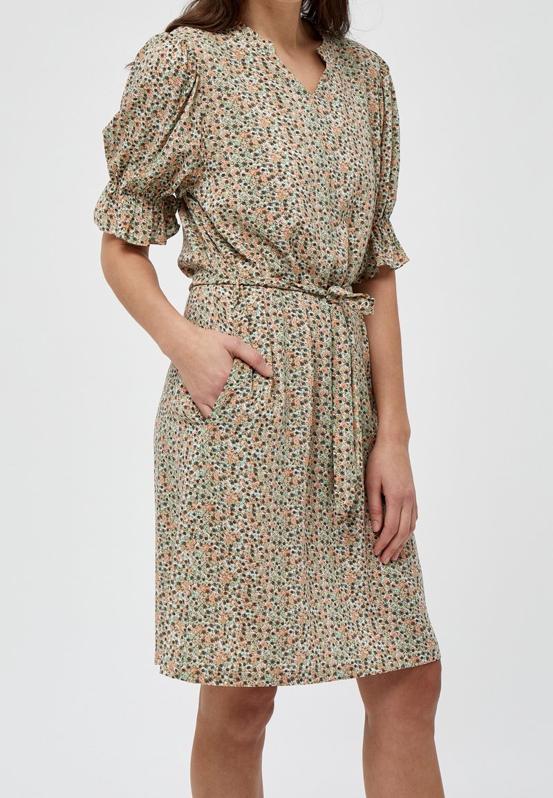Femme portant une robe fleurie à manches courtes avec une ceinture nouée à la taille et une main dans la poche, debout devant un fond uni.