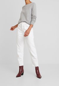 Szary sweter z dzianiny, białe jeansy o prostych nogawkach oraz bordowe teksturowane botki za kostkę. Sweter ma okrągły dekolt i ściągacze na mankietach.