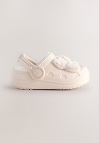 Lysebeige skumtræsko med en blød, pelslignende overdel, justerbar rem, tekstureret sål og dekorative pjuske-detaljer foran.