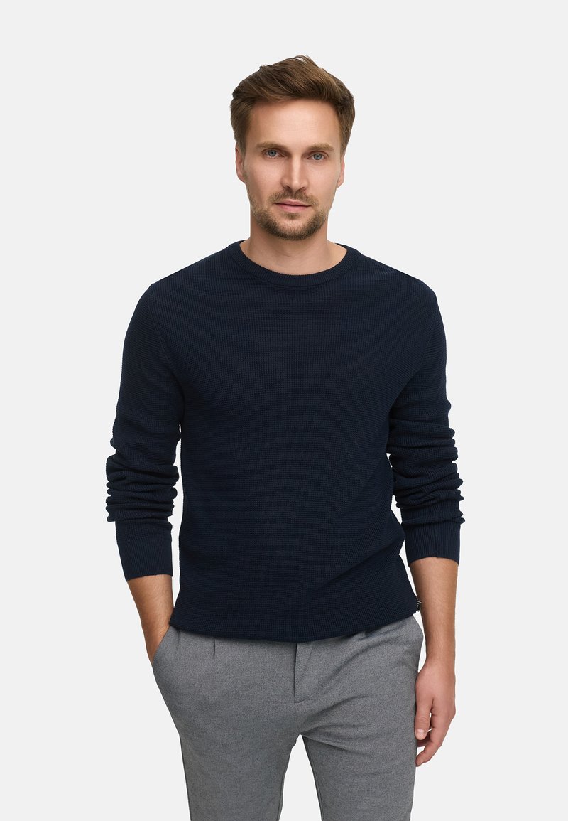 Marineblau gestrickter Pullover mit rundem Halsausschnitt und langen Ärmeln, kombiniert mit hellgrauen Hosen. Lockerer Schnitt mit gerippten Bündchen.