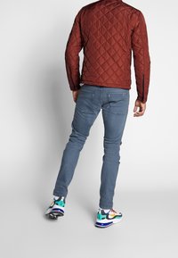 Gesteppte braune Jacke, hellblaue Jeans und mehrfarbige Turnschuhe. Die Jacke hat ein Rautenmuster, Reißverschlussdetails und ein figurbetontes Design.