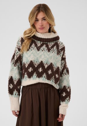 Femme portant un pull en grosse maille avec des motifs géométriques marron et bleu clair, une jupe marron et une expression neutre sur un fond uni.