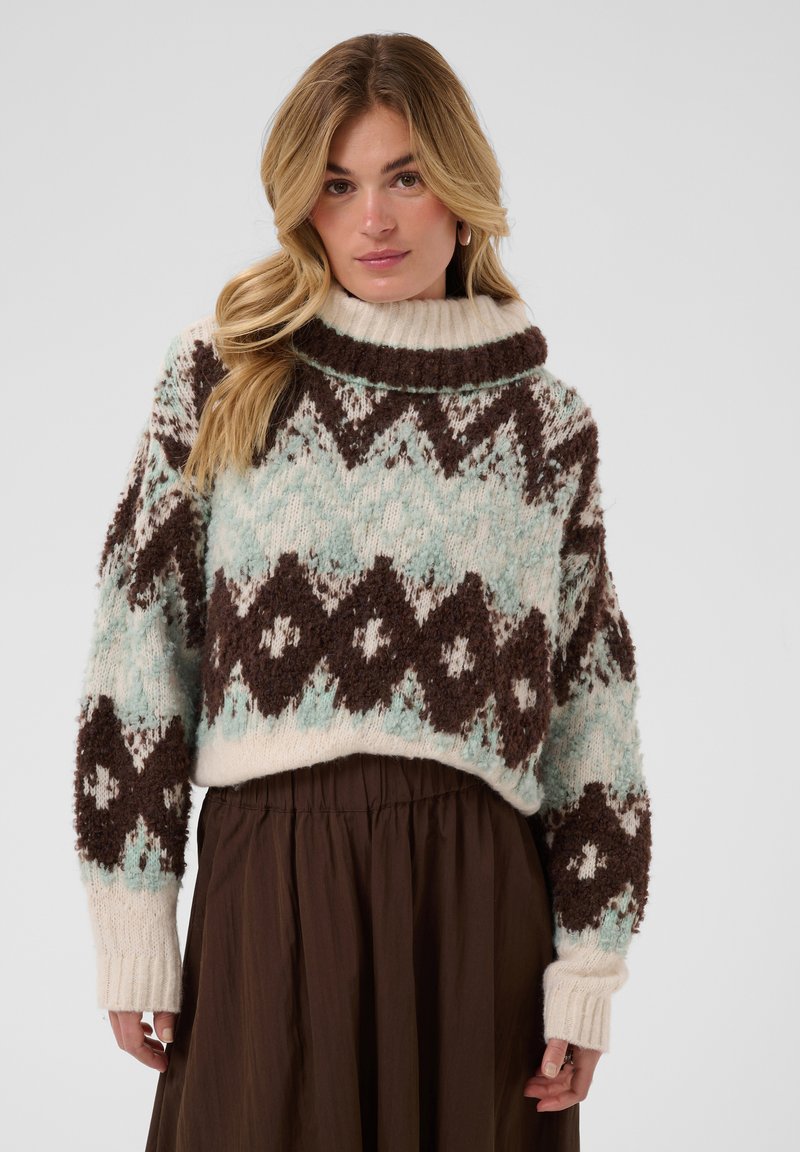 Femme portant un pull en grosse maille avec des motifs géométriques marron et bleu clair, une jupe marron et une expression neutre sur un fond uni.
