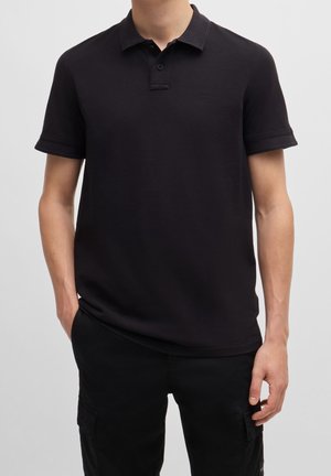 Schwarzes Poloshirt aus Baumwolle mit klassischem Kragen, kurzen Ärmeln und einem dezenten Logo auf der Brust. Glatte Textur und figurbetontes Design.