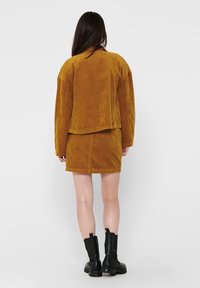 Completo giacca e gonna in velluto a coste di colore giallo senape, con texture a nervature verticali. Abbinato a stivali neri alla caviglia. Vista posteriore mostrata.