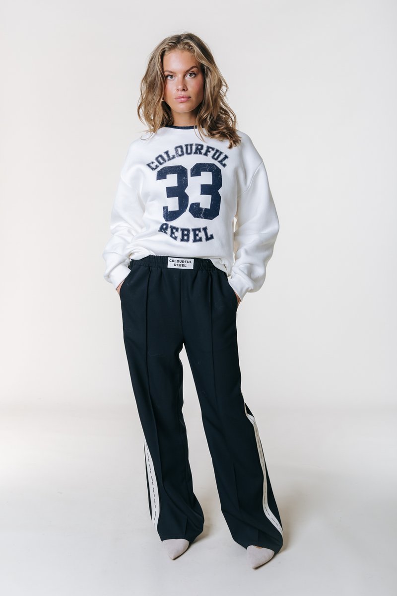 Λευκό φούτερ με μπλε σκούρα αριθμό "33" και κείμενο "COLOURFUL REBEL", συνδυασμένο με μαύρο παντελόνι που διαθέτει λευκές πλαϊνές ρίγες και υφασμένη υφή.
