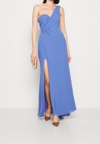 Asymetrické večerní šaty v těsném střihu v světle modré barvě s volně splývající tkaninou a rozparkem po stehno. Doplněné o hnědé stiletto lodičky a psaníčko.