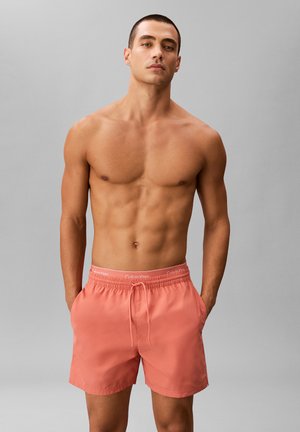Oberkörperfreier Mann mit kurzem Haar, der korallenpinke Calvin Klein-Badeshorts trägt und mit den Händen in den Taschen vor einem schlichten grauen Hintergrund steht.
