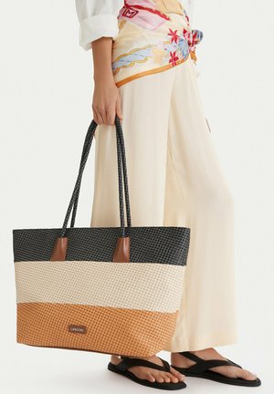 Persona vistiendo pantalones crema, cinturón bufanda floral y sandalias negras sosteniendo un gran bolso tejido tricolor con asas largas.