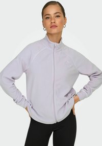 Donna con i capelli raccolti indossante orecchini a cerchio e una giacca in pile leggera di colore viola chiaro, con le mani sui fianchi, su sfondo bianco.