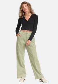 Black cropped cardigan with long sleeves, paired with light green corduroy wide-leg pants. Model stands casually with hands in pockets.