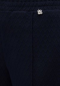 Tissu bleu marine foncé avec motif matelassé en losange et petite étiquette logo argentée « HUGO » près de la couture.