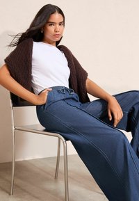 A széles szárú farmernadrág kék árnyalatban, húzózsinóros derékrésszel készült. Barna kötött kardigán van fehér póló fölött viselve. A modell ül.