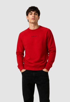 Giovane uomo con capelli ricci scuri che indossa una felpa rossa con la scritta "Goudi Jeans", pantaloni neri, in piedi con le mani in tasca su sfondo grigio.