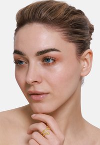 Bague en or en forme de serpent sur une main à la peau lisse, encadrée par un visage aux contours doux, avec un maquillage léger et des cheveux soigneusement coiffés.