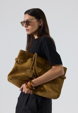 Mujer con cabello largo y oscuro que lleva gafas de sol y un conjunto negro sostiene un gran bolso de ante marrón y pulseras de ámbar sobre un fondo gris.