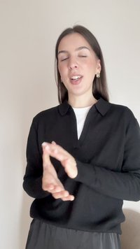 Mlada žena s dugom smeđom kosom, odjevena u crnu majicu i zlatne naušnice, gestikulira rukama i govori sa zatvorenim očima na jednobojnoj pozadini.