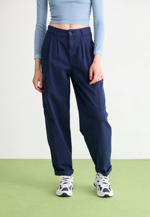 Henkilö, jolla on päällään tummansininen taskuhousut, joissa mansetit, valkoiset ja tummansiniset lenkkarit sekä vaaleansininen pitkähiha cropped-top, seisoo vihreällä lattialla.