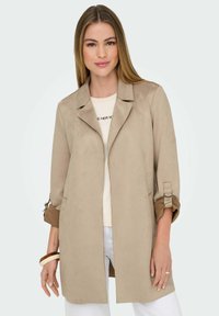 Donna che indossa un lungo giubbotto beige con maniche arrotolate sopra una camicia e pantaloni bianchi, in piedi contro uno sfondo chiaro e uniforme.