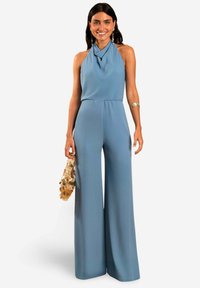 Combinaison bleu clair avec un col halter drapé et un design à jambes larges. Caractéristiques d'un tissu lisse et d'une taille cintrée. Les accessoires comprennent un sac texturé.