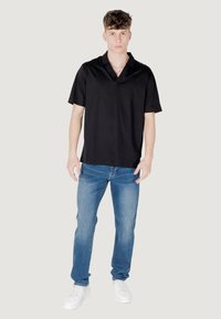 Polo shirt nero a maniche corte con colletto, abbinato a jeans di denim azzurro chiaro e sneakers bianche, in piedi su uno sfondo neutro.