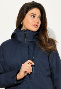 Dunkelblauer Winterjacke mit hohem Kragen, verstellbarer Kapuze und Reißverschlusstaschen. Verfügt über einen strukturierten Stoff und Druckknopfmanschetten.