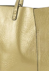 Close-up van een getextureerde gouden leren tote bag met gestikte handvatten en metalen knoopdetails.